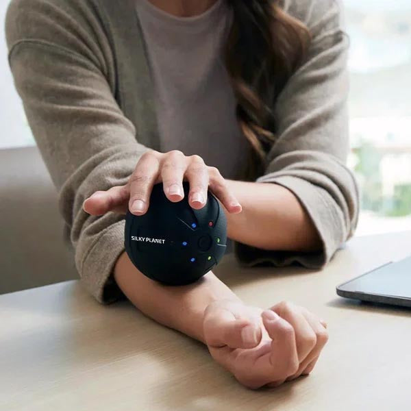 En person använder Silky Planets massageboll på underarmen vid ett skrivbord.